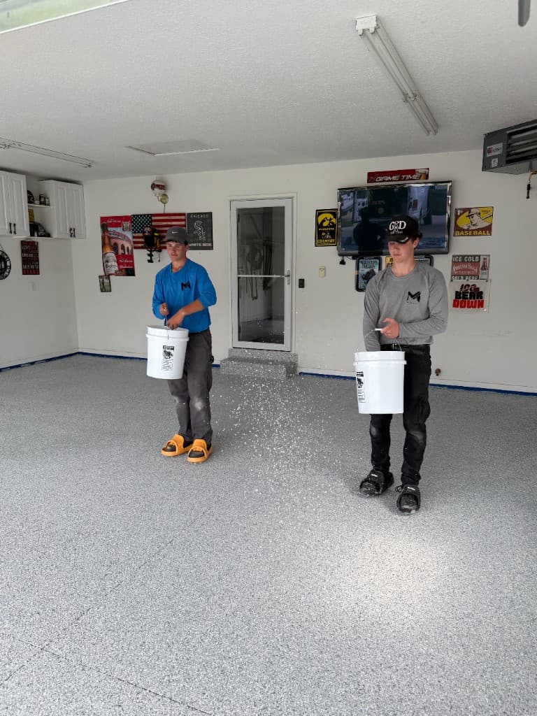 Two M-Pact installers in branded shirts broadcasting full decorative flake onto a wet garage floor coating.